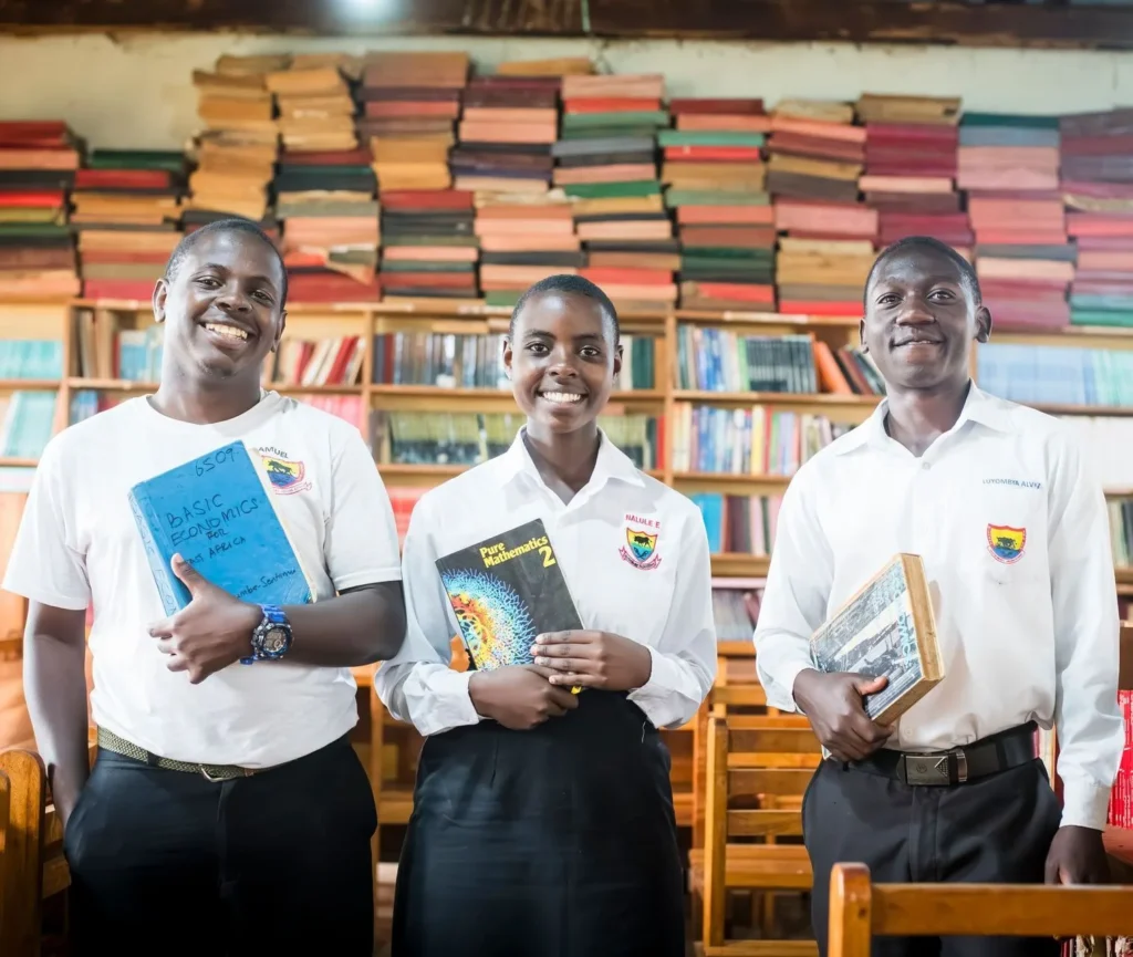 Three students holding textbooks in front of shelves of books in a school library